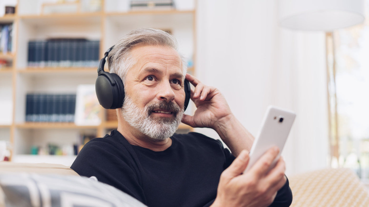 Älterer Mann hört mit Kopfhörern Musik und schaut entspannt auf sein Smartphone als Symbol für aktives und gesundes Altern.